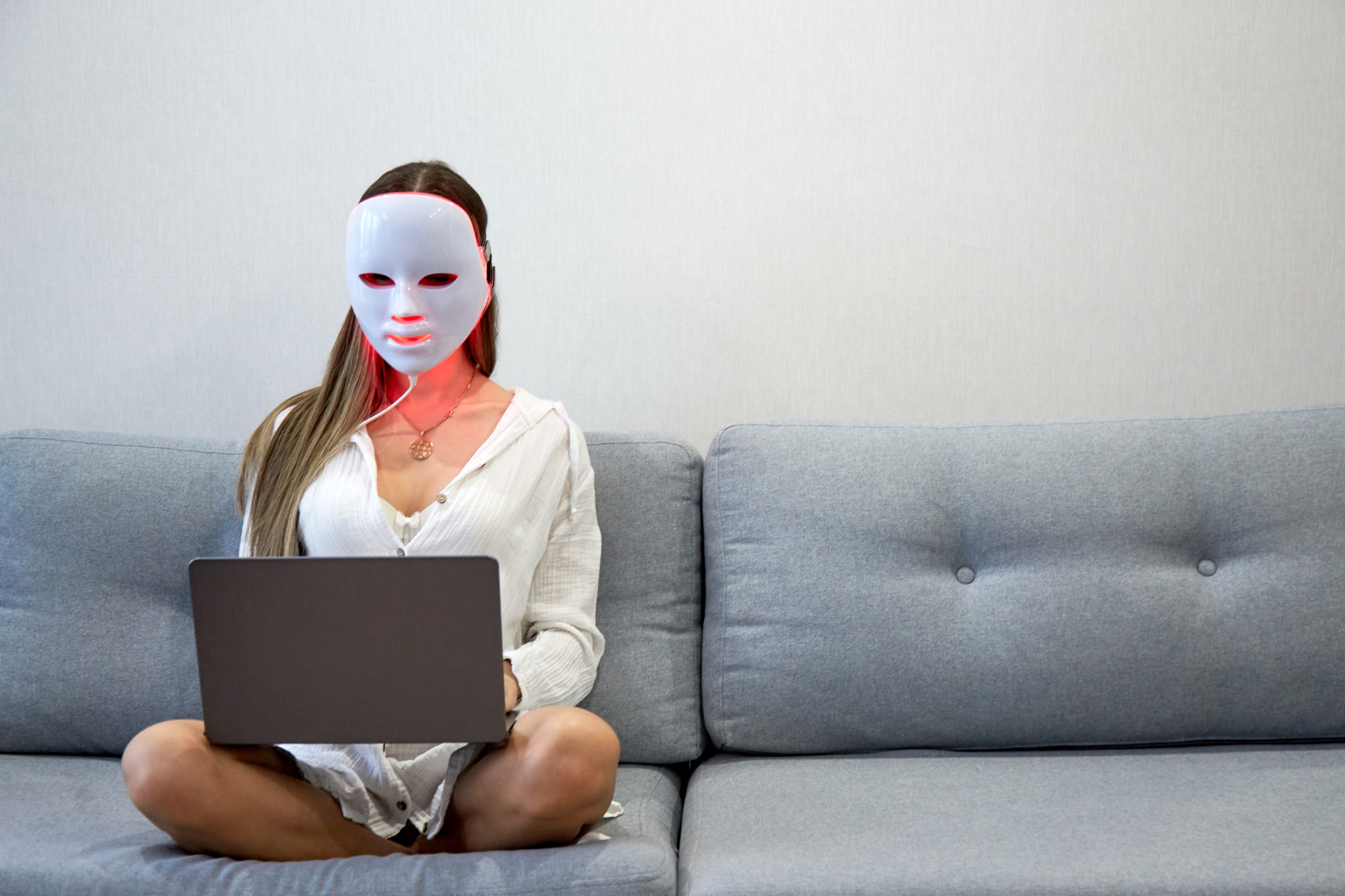 A beautiful girl with an LED mask on her head works at home on a laptop. Home skin care concept.